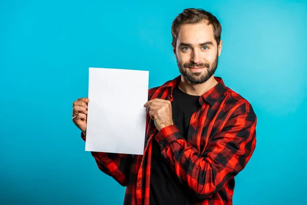 Красивий чоловік тримає вертикальний білий паперовий плакат a4. Копіювати простір. Усміхнений хіпстер у червоній сорочці на синьому фоні . — стокове фото
