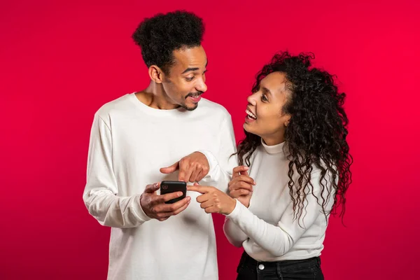 Young couple using mobile device. Man talking about apps and explains to girl how to use application. Red studio background