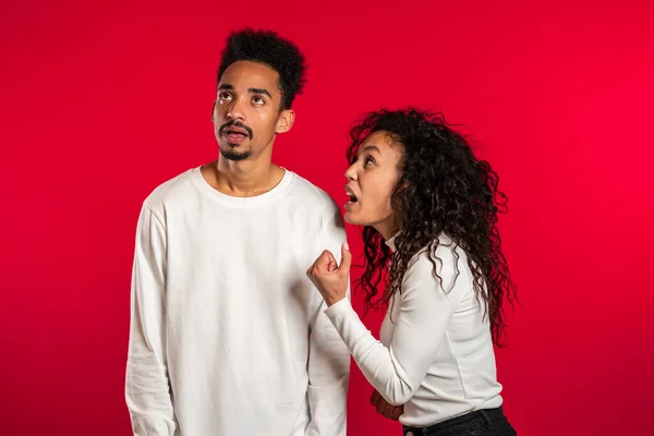 Young african woman emotionally screaming at her husband or boyfriend on red background in studio. Bored man rolling his eyes. Concept of conflict, problems in relationships. — Stock Photo, Image