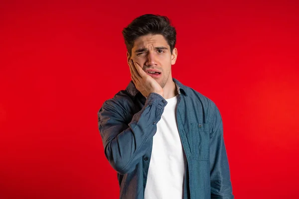 Jeune homme beau en chemise denim avec douleur dentaire sur fond de studio rouge. Maux de dents, problèmes dentaires, stomatologie et concept de médecine . — Photo