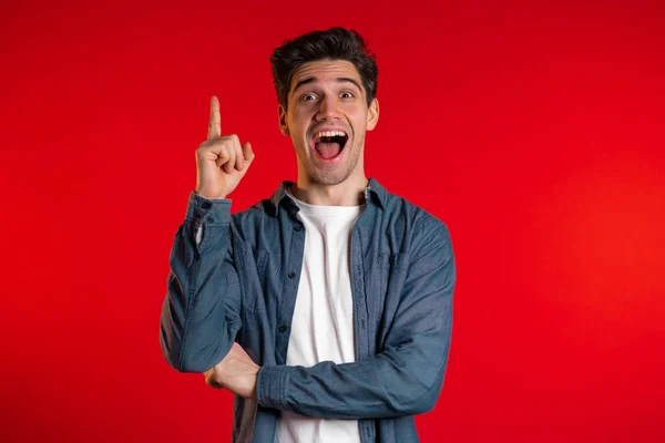 Portrait of young thinking pondering man having idea moment pointing finger up on red studio background. Smiling happy guy showing eureka gesture. — Stock Photo, Image