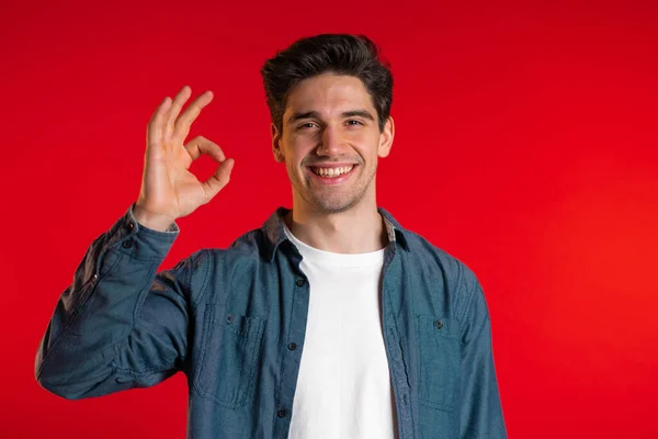 Young handsome man making OK sign over red background and smiles to camera. — Stock Photo, Image
