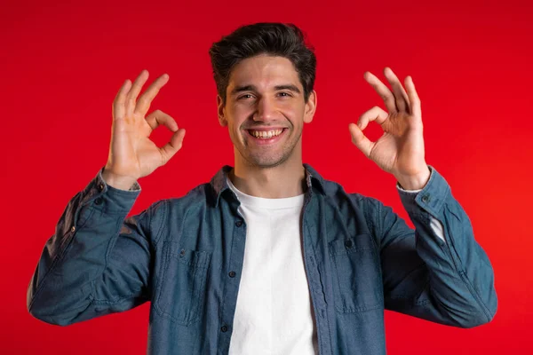 Young handsome man making OK sign over red background and smiles to camera. — Stock Photo, Image