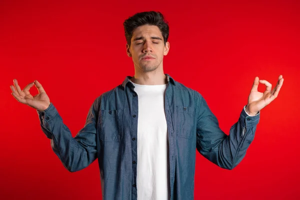 Calm student guy or businessman relaxing, meditating. Handsome european man calms down, breathes deeply with mudra om on red background — Stock Photo, Image