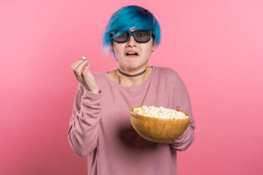 Üç boyutlu gözlüklü genç bir kadın korku filmi izliyor ve pembe stüdyo arka planında patlamış mısır yiyor. Filmdeki beklenmeyen entrika yüzünden mavi saçlı ürkek bir kız..