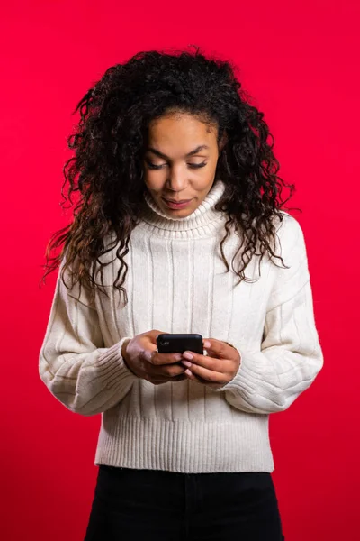 Chica con el pelo afro utilizando el teléfono inteligente, navegar por Internet o jugar a juego en el fondo del estudio rojo. Tecnología moderna - aplicaciones, redes sociales — Foto de Stock
