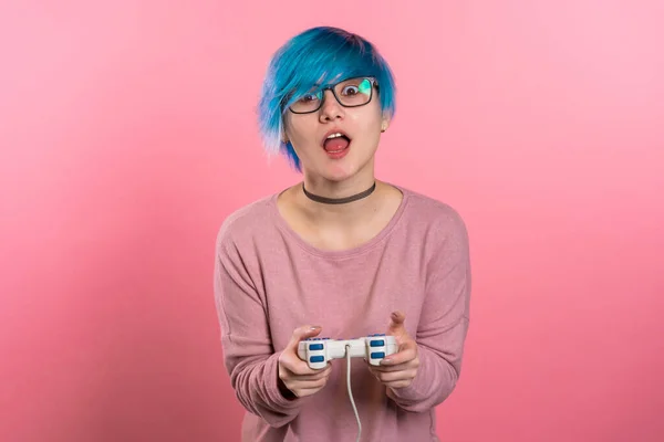 Hübsches junges Mädchen mit blauer Frisur beim Videospiel auf dem Fernseher mit Steuerknüppel an der rosafarbenen Studiowand. Einsatz moderner Technologie. — Stockfoto