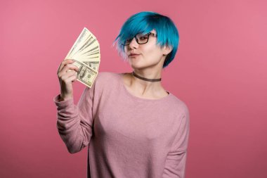 Tatmin olmuş mavi saçlı, heyecanlı kız para gösteriyor. Pembe duvarda Amerikan doları banknotları. Başarı, kazanç, zafer sembolü.