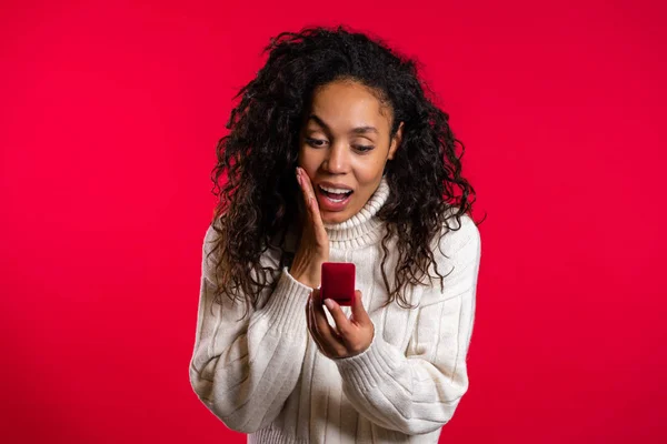 Amazed african american young woman holding small jewelry box with proposal diamond ring on red wall background. Girl smiling, she is happy to get present, proposition for marriage. — Stock Photo, Image