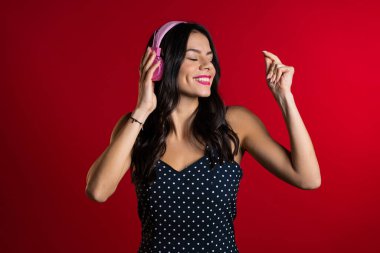 Uzun saçlı, neşeli, gülümseyen, kırmızı arka planda pembe kulaklıklarla dans eden güzel bir genç kız. Müzik, dans, radyo konsepti.
