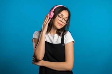 Uzun saçlı güzel Asyalı bir kız. Eğleniyor, gülümsüyor, kulaklıklarla dans ediyor. Stüdyoda mavi arka planda. Müzik, dans, radyo konsepti,