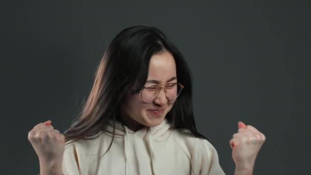 Азійська дівчина з довгим волоссям дуже задоволена і щаслива, вона показує "так" жест перемоги, вона досягла результату, цілей. Здивована щаслива жінка на сірому фоні — стокове відео