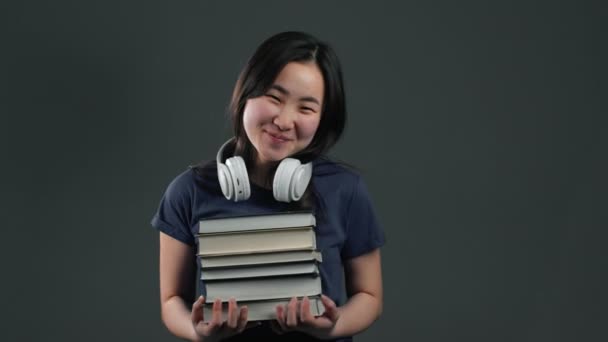 Asiatisk student flicka med hörlurar på grå bakgrund i studio rymmer stack med universitetsböcker från biblioteket. Kvinna ler, hon är glad att ta examen. — Stockvideo