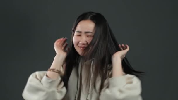 Engraçado mulher asiática com cabelos longos se divertindo, sorrindo, dançando em estúdio contra fundo cinza. Música, conceito de dança, câmera lenta — Vídeo de Stock
