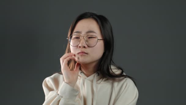 Thinking woman looking up and around on grey background. Worried contemplative face expressions. Pretty asian girl model in white hoodie — Stock Video