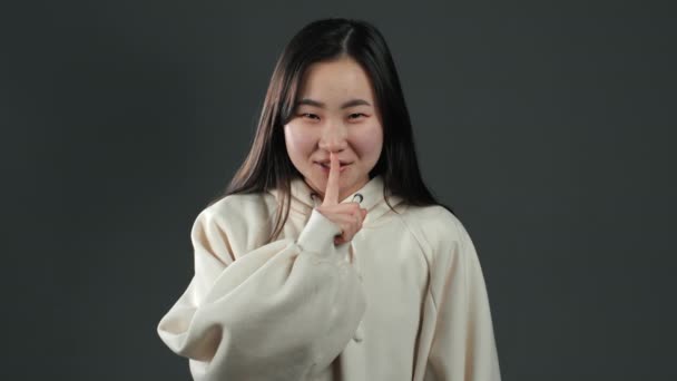 Leende asiatisk kvinna med långt hår håller fingret på läpparna över grå bakgrund. Gest av shhh, hemlighet, tystnad. Närbild. — Stockvideo