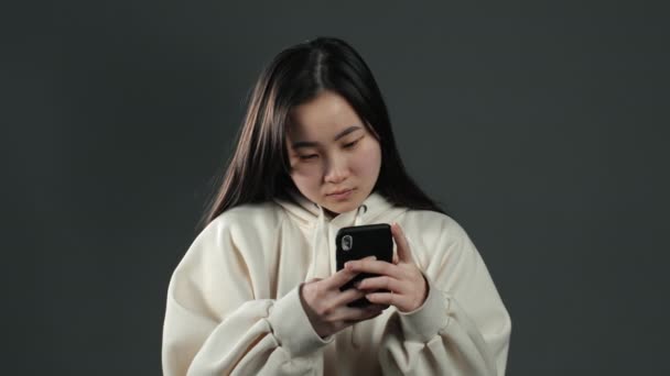 Menina asiática sms mensagens de texto, usando o aplicativo no smartphone. Mulher bonita navegando na internet com telefone celular. Imagens de estúdio cinza . — Vídeo de Stock