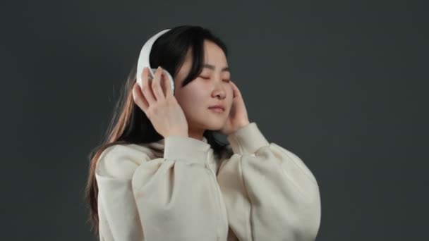 Menina coreana muito jovem com cabelos longos sorrindo, dançando e desfrutando de música com fones de ouvido em estúdio contra fundo cinza. Música, dança, conceito de rádio . — Vídeo de Stock