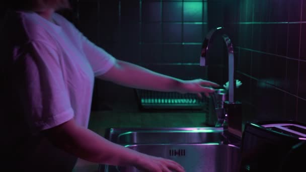 Vrouw opent kraan met water in moderne keuken met neon licht en vuil glas wassen. — Stockvideo