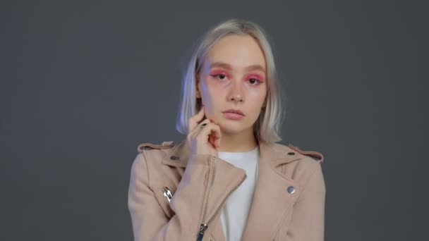 Serious hipster woman isolated on grey studio background. Portrait of tired lady with platinum blonde hair in pink jacket looking to camera. — Stock Video