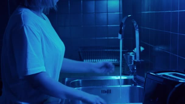 Vrouw opent kraan met water in moderne keuken met neon licht en vuil glas wassen. — Stockvideo