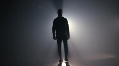 Deri ceketli genç adam dumanlı stüdyoda ya da kamera önünde yürüyor. Arka planda ışık, parlak sahne ışığı