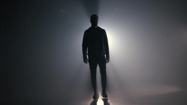 Hombre joven en chaqueta de cuero está caminando a través de un estudio ahumado o escena directamente a la cámara. Retroiluminación, proyector brillante en el fondo — Vídeo de stock