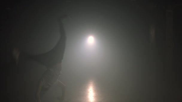 Güçlü bir yetişkin adam sahne ışıkları altında dumanlı karanlık stüdyoda inanılmaz numaralar sergiliyor. Hile, parkur, atlama pratiği konsepti. Yavaş çekim. — Stok video