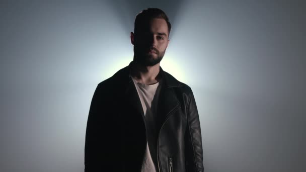 Retrato dramático de un joven europeo con barba en chaqueta de cuero mirando a la cámara. Estudio de humo oscuro o escenario. Retroiluminación, proyector brillante en el fondo — Vídeos de Stock