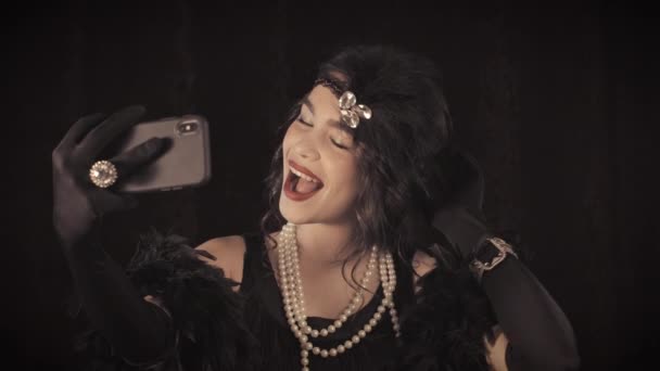 Kvinna klädd i Gatsby stil med fjädrar boa gör selfie med smartphone på mörk bakgrund. Roaring tjugoårsåldern, retro, fest, mode koncept. Sepia retrofilmseffekt — Stockvideo