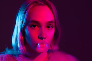 Alışılmadık bir yüzü olan genç ve güzel bir kız gece şehrin parlak pembe neon ışıklarıyla çiğneme baloncuğunu şişiriyor. Boyalı menekşe saç. Yeniyetme ergen konsepti.