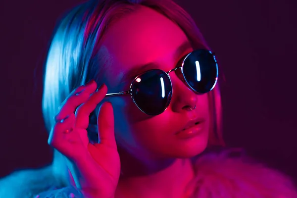 Close up portrait of millennial pretty glamour girl in fluffy fur coat in neon light. Dyed blue and pink hair. Mysterious hipster teenager in glasses. — Stock Photo, Image