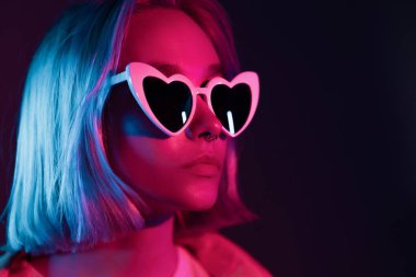 Stüdyo arka planında güneş gözlüklü, alışılagelmedik güzellikteki göz kamaştırıcı ve moda kızı portresi. Pembe ve mor neon ışıklı genç kadın.. 