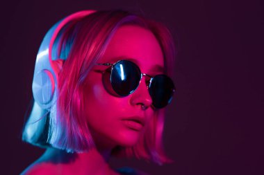 Boyalı saçları ve güneş gözlükleriyle kulaklıkla müzik dinleyen ve neon ışıklı arka planda şarkı söyleyen muhteşem bir bayan. Büyüleyici hippi kız gözleri kapalı dans ediyor..