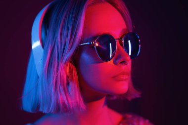 Boyalı saçları ve güneş gözlükleriyle kulaklıkla müzik dinleyen ve neon ışıklı arka planda şarkı söyleyen muhteşem bir bayan. Büyüleyici hippi kız gözleri kapalı dans ediyor..