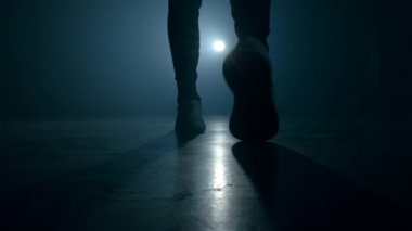 Spor ayakkabılı genç adam bacakları silueti dumanlı stüdyoda ya da kamera önünde yürüyor. Arka planda ışık, parlak sahne ışığı.
