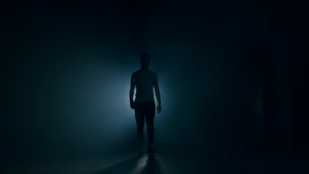 Young man in sports wear walking through smoky studio or scene directly to camera. Backlight, bright spotlight on background. — Stock Video