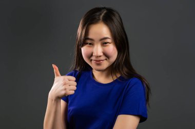 Asyalı kadın onaylama jesti yapıyor. Kazandım. Başarı. Gri arka plandaki olumlu genç kız kameraya gülümsüyor ve baş parmağını kaldırıyor.