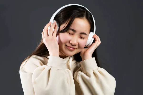 Netter asiatischer Teenager, der mit Kopfhörern Musik hört. Porträt eines tausendjährigen hübschen Mädchens mit langer Frisur auf grauem Hintergrund — Stockfoto