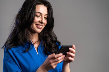 Olgun kadın mesajlaşıyor, akıllı telefondaki uygulamayı kullanıyor. İnternette cep telefonuyla sörf yapan güzel bir kadın. Gri stüdyo portresi.