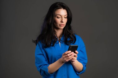Olgun kadın mesajlaşıyor, akıllı telefondaki uygulamayı kullanıyor. İnternette cep telefonuyla sörf yapan güzel bir kadın. Gri stüdyo portresi.