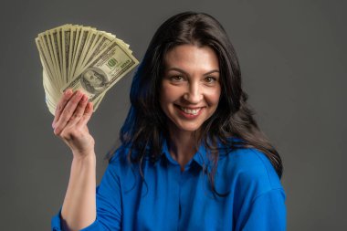 Gri duvarda Amerikan doları banknotları gösteren mutlu ve heyecanlı yetişkin kadın. Başarı, kazanç, zafer sembolü.