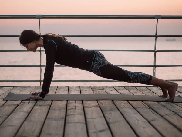 Ung brunett kvinna gör yoga övningar på trä havet banvallen på morgonen. Flicka i svart sport kostym. Stretching, övning, hälsosam livsstil Concept — Stockfoto