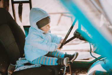 Sevimli küçük bebek eski eski bir arabada oturuyor ve elinde merakla tekerleği tutuyor. Çocuk, otomobil, çocukluk hayalleri, aile ve aşk kavramı.