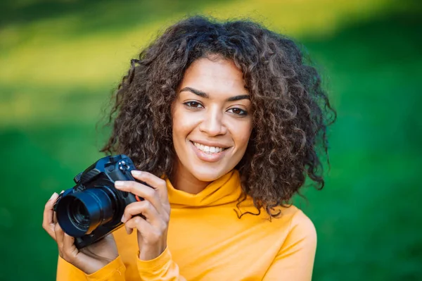 黄色の若いかなりアフリカの女性は、緑の背景にデジタル一眼レフカメラで写真を撮る。女の子は写真家として楽しんで笑っている. — ストック写真