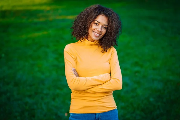 Portret afrykańskiej dziewczyny w zielonym parku. Młoda kobieta z kręconą fryzurą w żółtym stroju cieszy się przyrodą. — Zdjęcie stockowe