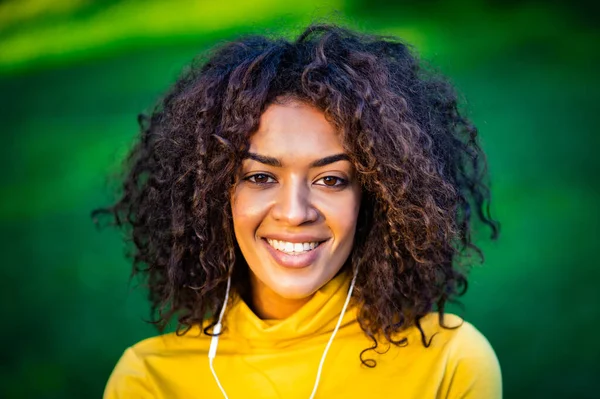 Портрет афро-американської дівчини в білих навушниках. Молода жінка з кучерявою зачіскою в жовтому кольорі любить музику в парку.. — стокове фото