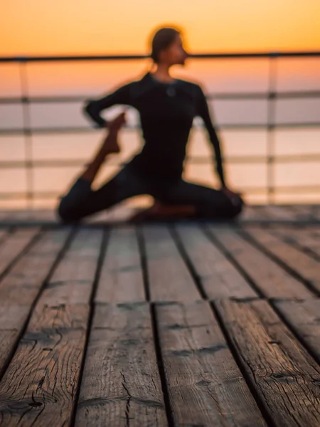 Jeune femme floue faisant des exercices de yoga sur un remblai en bois le matin. Fille en costume de sport noir. Étirement, pratique, mode de vie sain concept — Photo