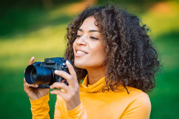 黄色の若いかなりアフリカの女性は、緑の背景にデジタル一眼レフカメラで写真を撮る。女の子は写真家として楽しんで笑っている. — ストック写真