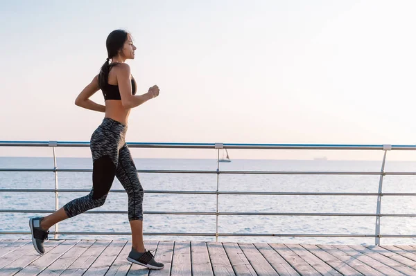 Sportowa młoda kobieta jogging rano przez morze drewniany nasyp. Sylwetka dziewczyny w stroju sportowym. Piękne światło słoneczne. Koncepcja zdrowego stylu życia. Przestrzeń kopiowania — Zdjęcie stockowe